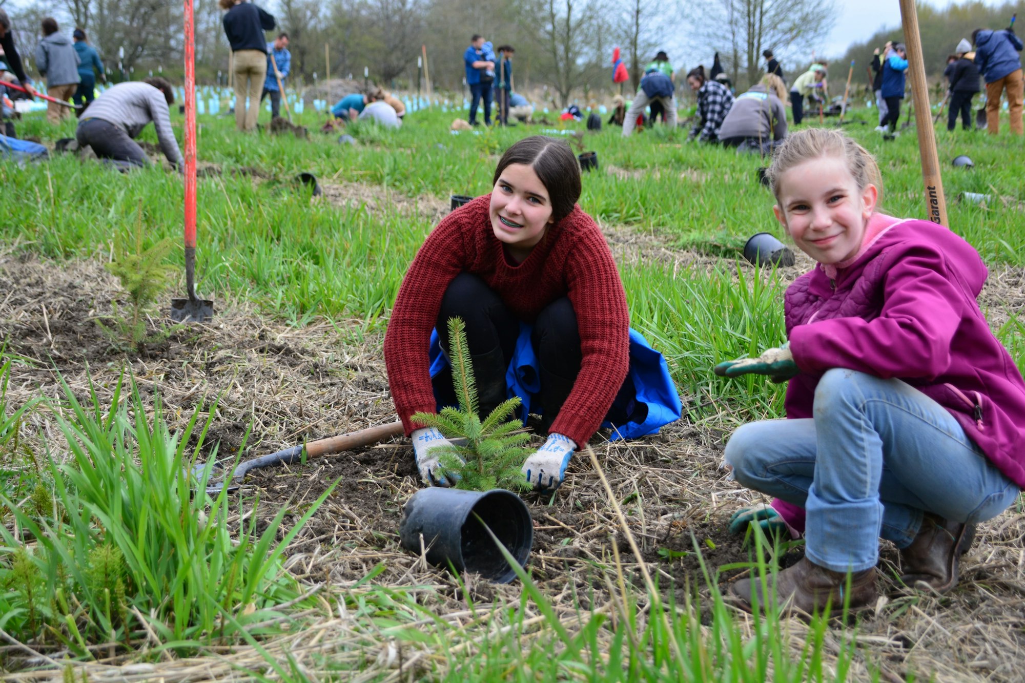 People planting trees in a field, with two young girls smiling at the camera.