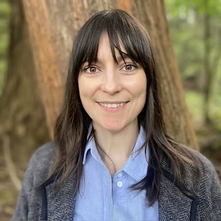 A person with long brown hair and a blue shirt smiles outdoors in front of a tree.