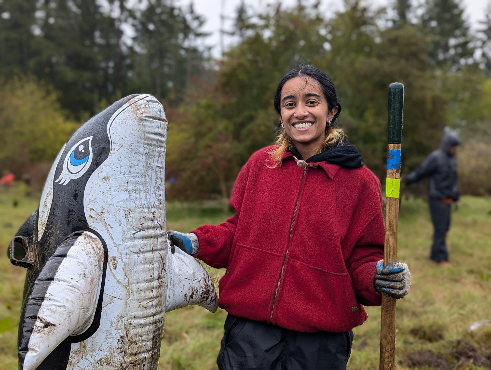 Person smiling, holding a shovel next to a large inflatable orca in a grassy area.