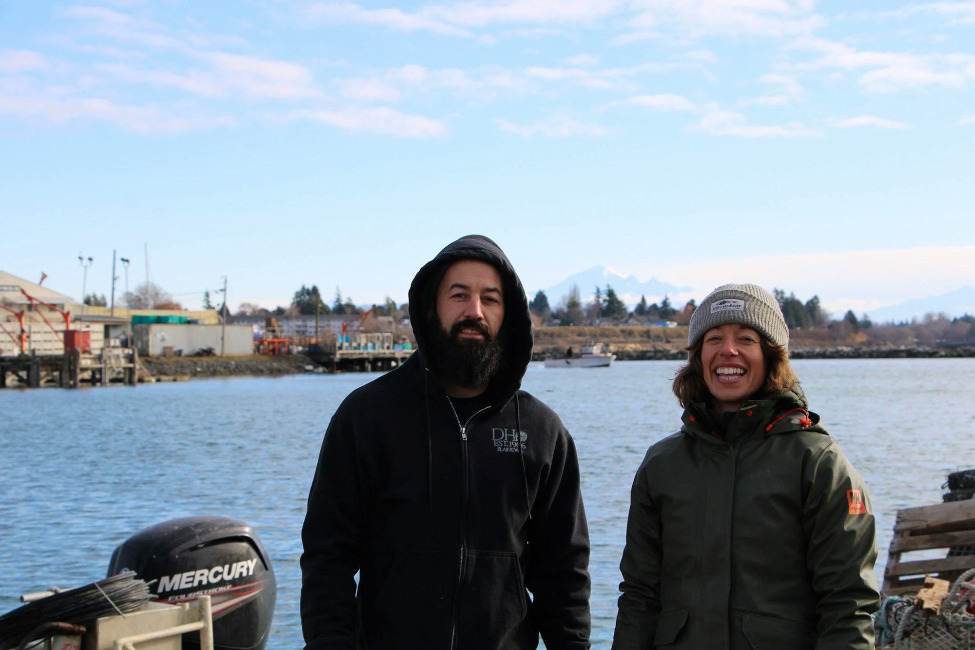 Two people in outdoor clothing smile by a waterfront, with a marina in the background.