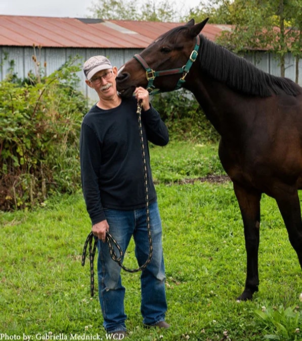 A man in a cap holding a horse's lead as the horse nuzzles his face in a grassy area.