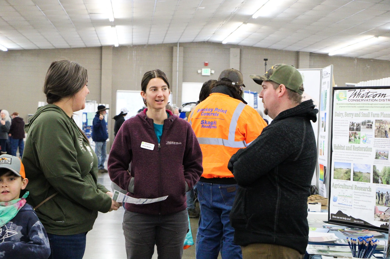 People are conversing at a public event with informational displays, including a Whatcom Conservation District booth.