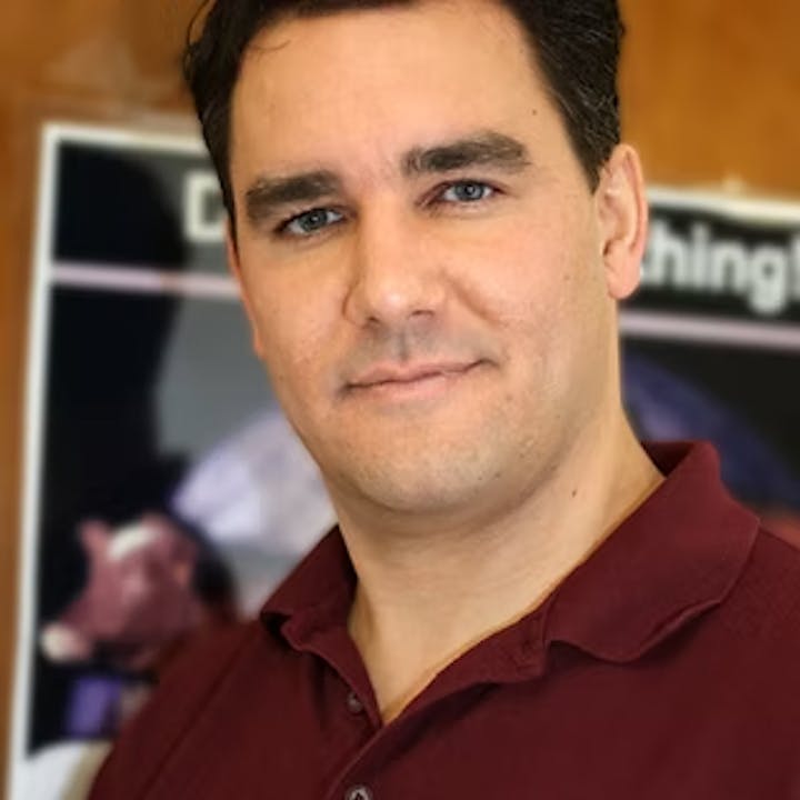 A man in a red shirt standing in front of a blurred poster featuring a cow.