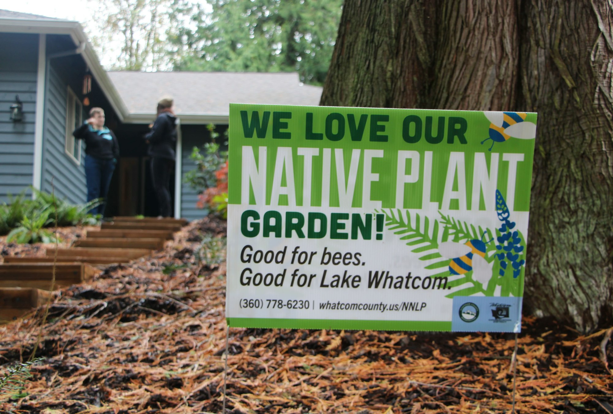 A sign reads, "We love our native plant garden! Good for bees. Good for Lake Whatcom," in front of a house and two people.