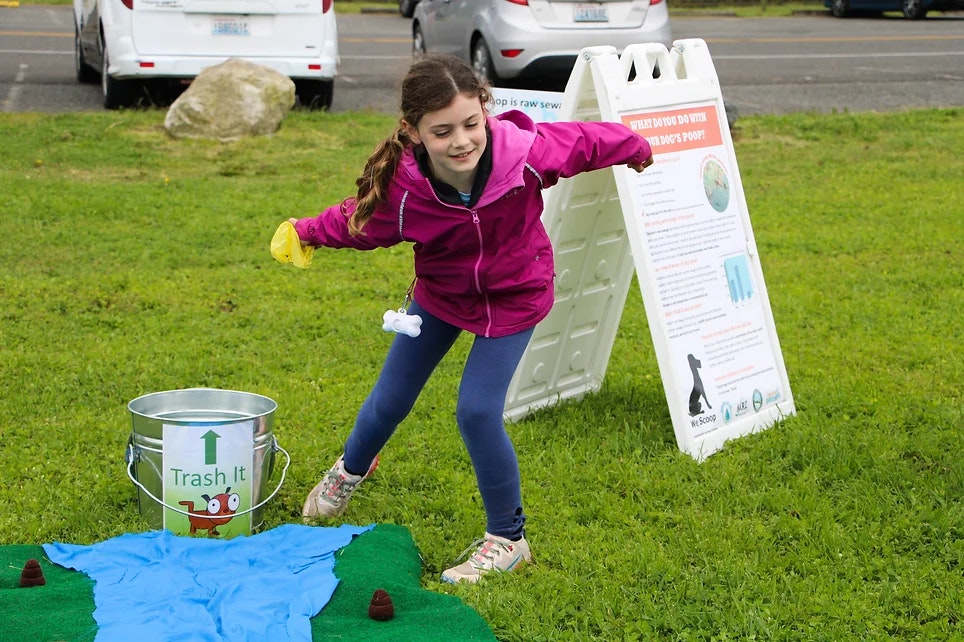 A child in a pink jacket plays a cleanup-themed game, aiming at a bucket labeled "Trash It" outdoors.