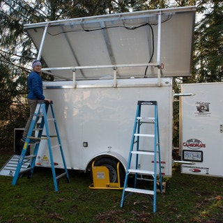 A person on a ladder is working on a trailer with a raised solar panel. Two ladders are positioned next to the trailer.