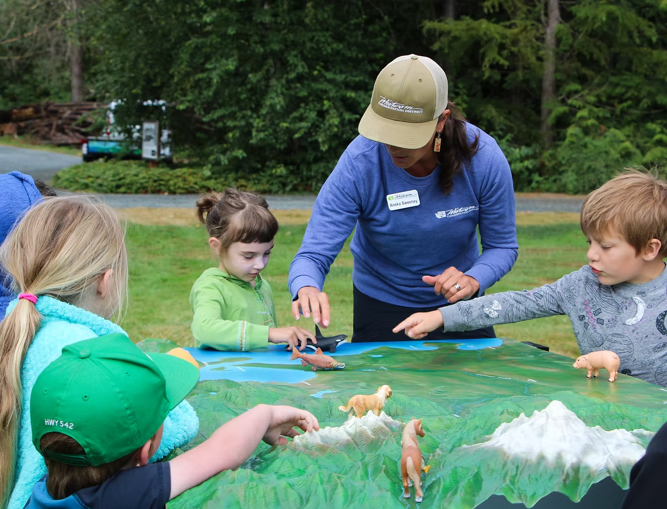 A woman and children interact with a nature-themed diorama outdoors.