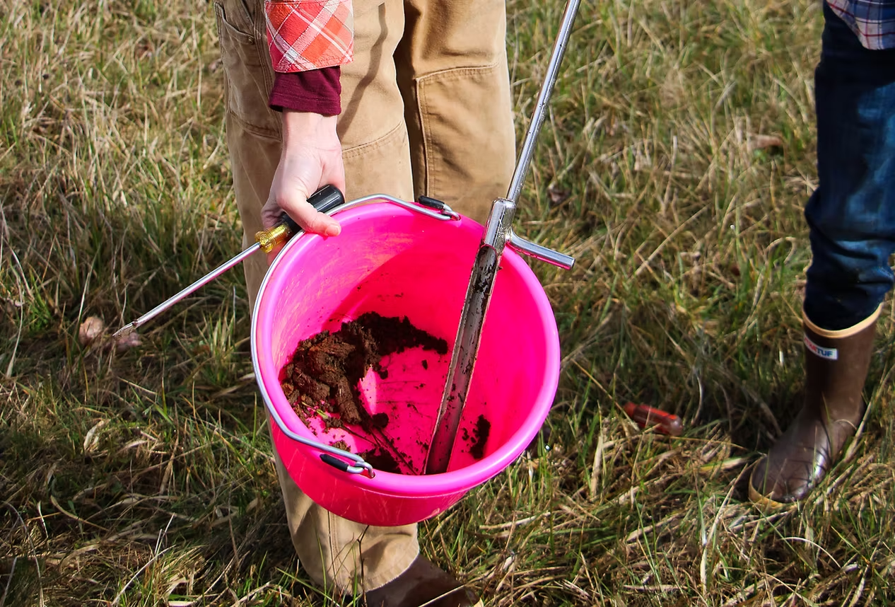A person holds a pink bucket containing soil samples and a soil probe, standing on grassy ground.