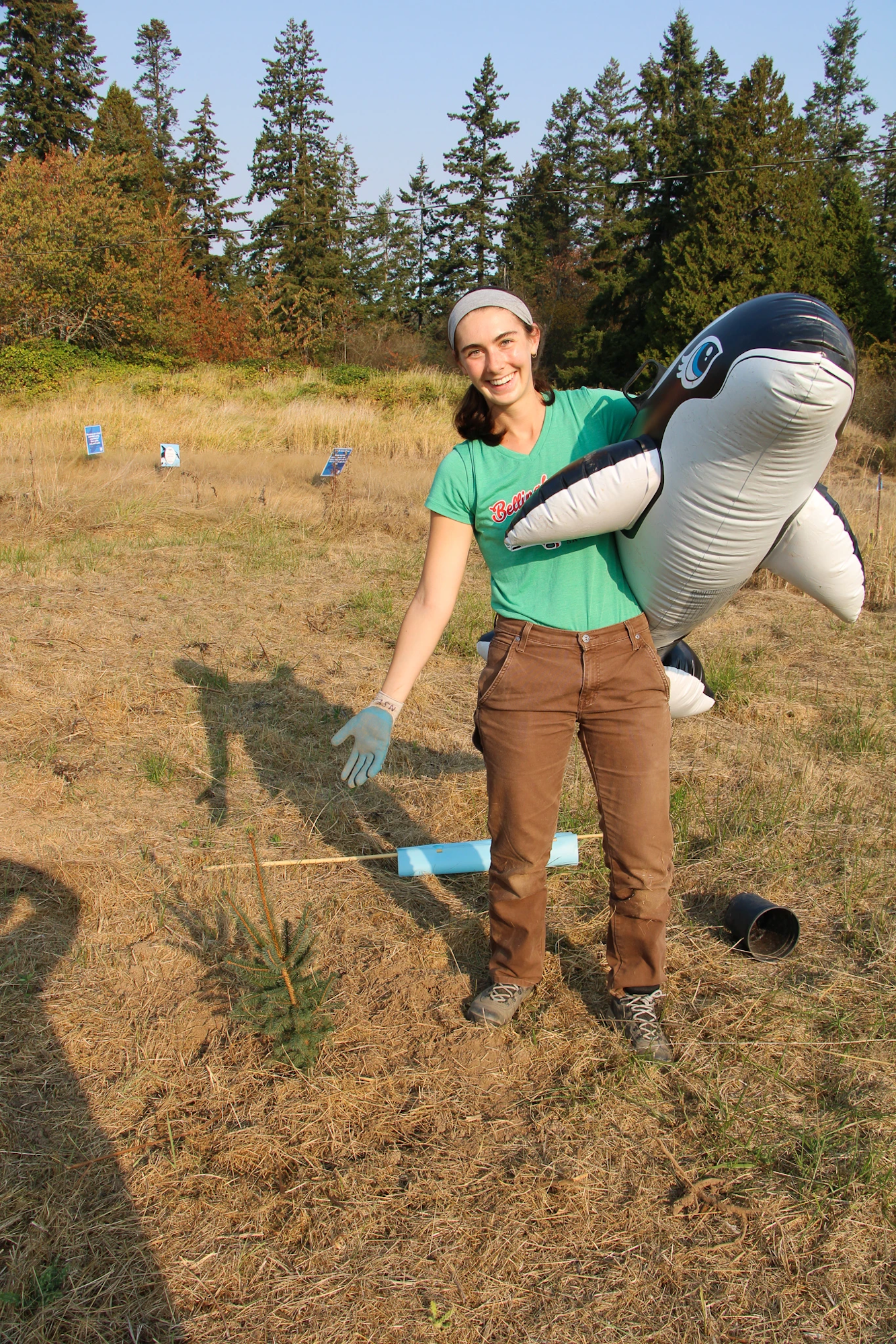 A person in a field holding an inflatable orca, wearing gloves, with trees and signs in the background.