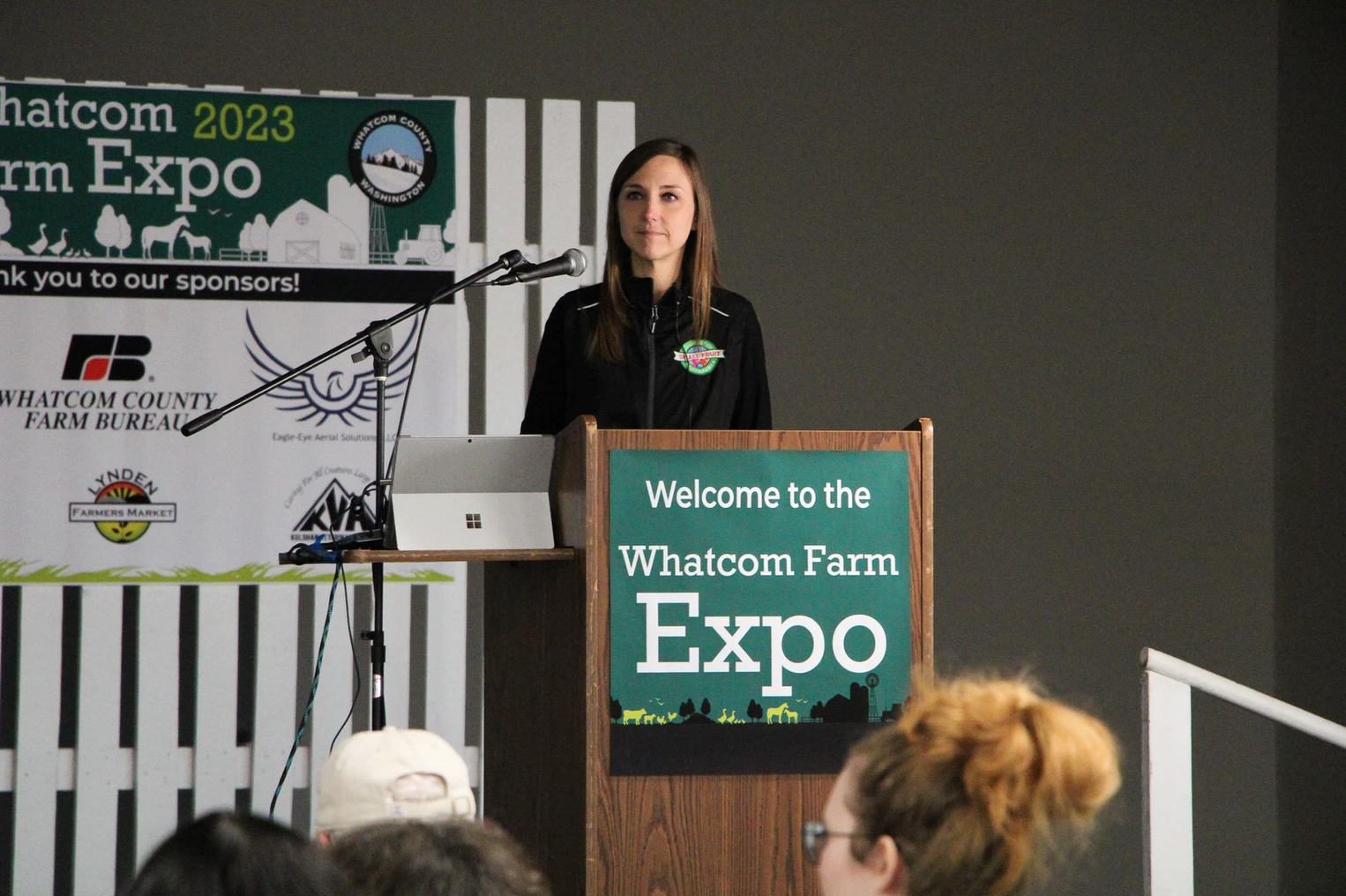 A speaker at a podium at the Whatcom Farm Expo, with a banner and sponsors in the background.