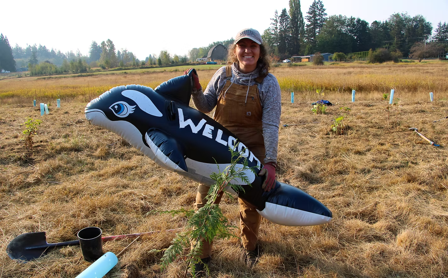 Person holding an inflatable orca with "WELCOME" written on it, in a field with tools and small plants.