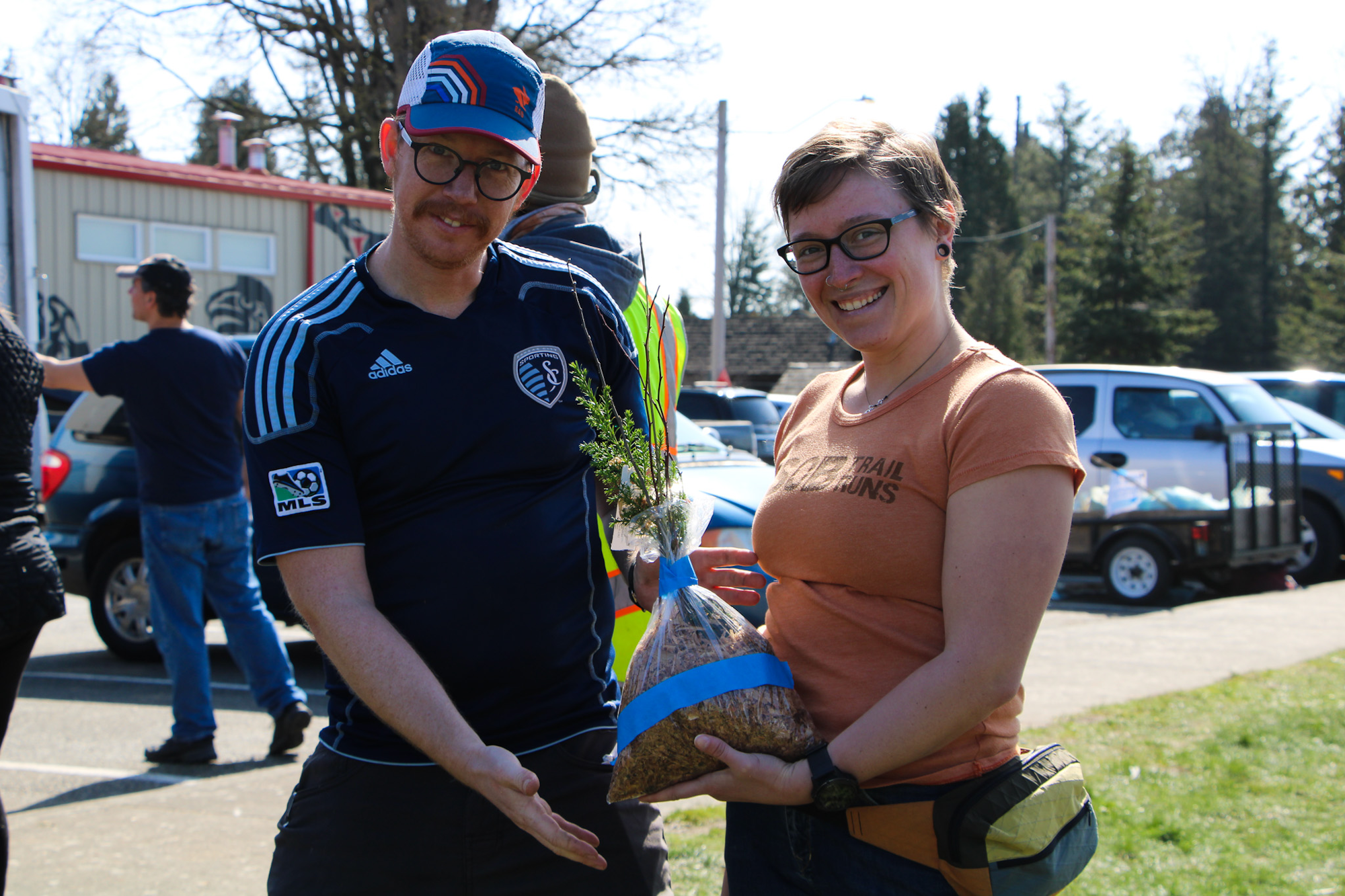 Two people smiling, holding a small tree in a bag of soil.