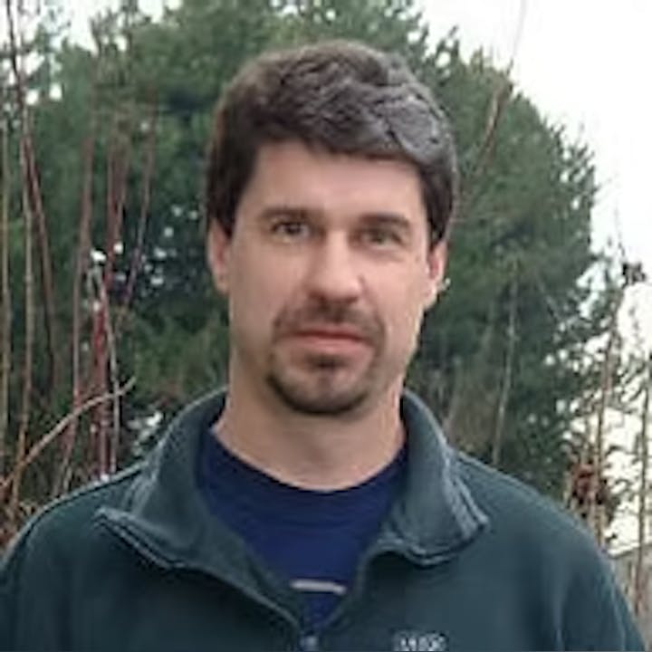 A man with dark hair and a beard, wearing a dark jacket, is standing outdoors with trees in the background.
