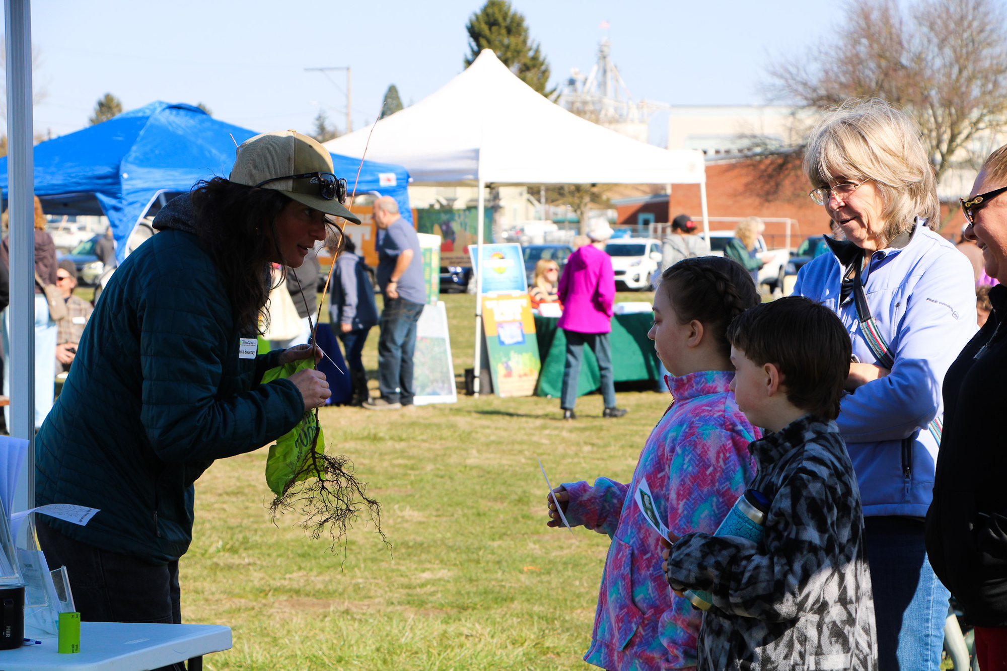 A person talks to a group at an outdoor event, holding a plant with roots. Tents and tables are set up in the background.