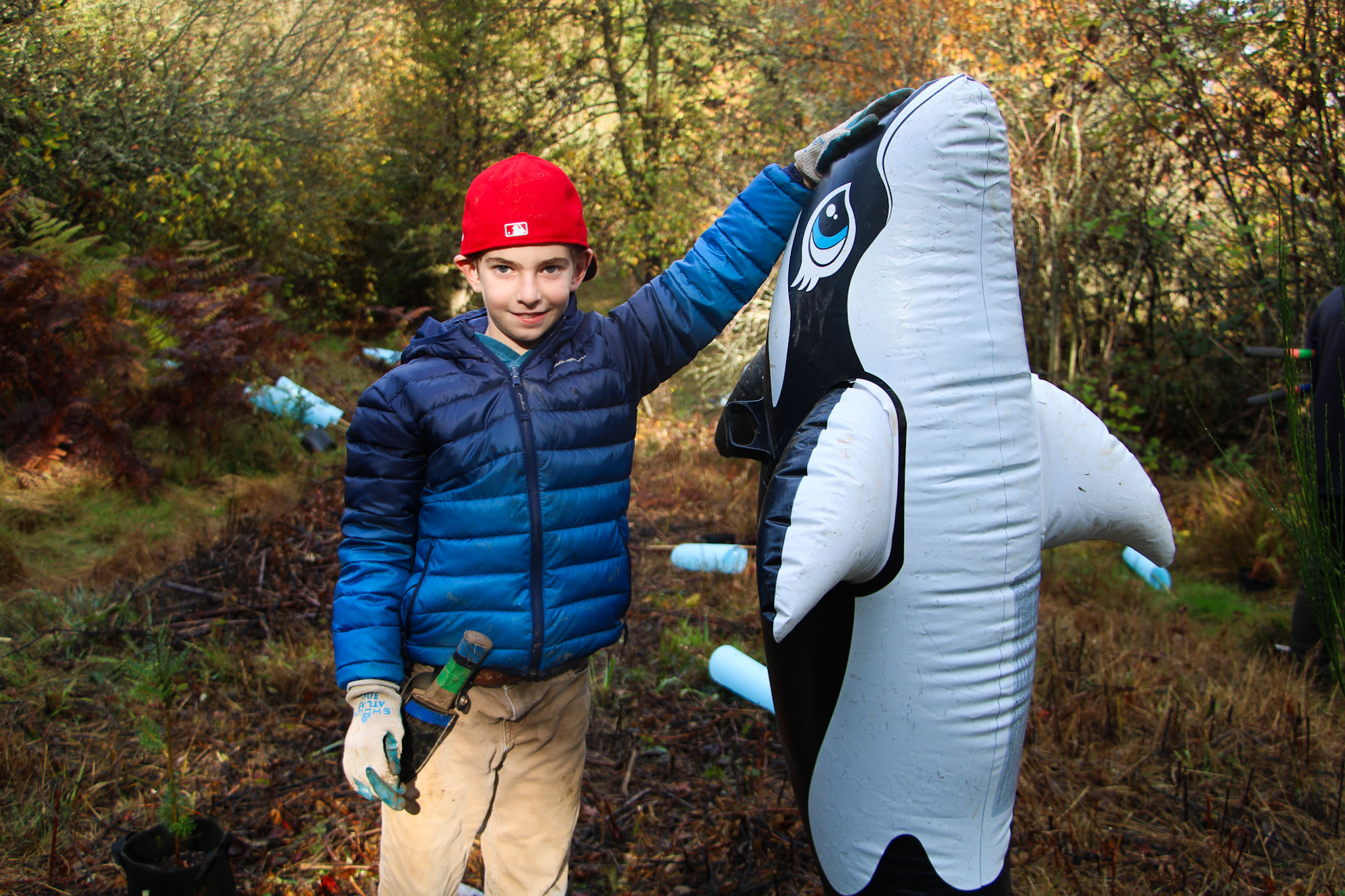 A person in a red cap and blue jacket, outdoors, stands with an inflatable orca and holds gardening tools.