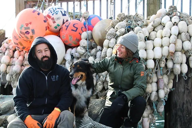 Two people with a dog, sitting in front of fishing nets and buoys.