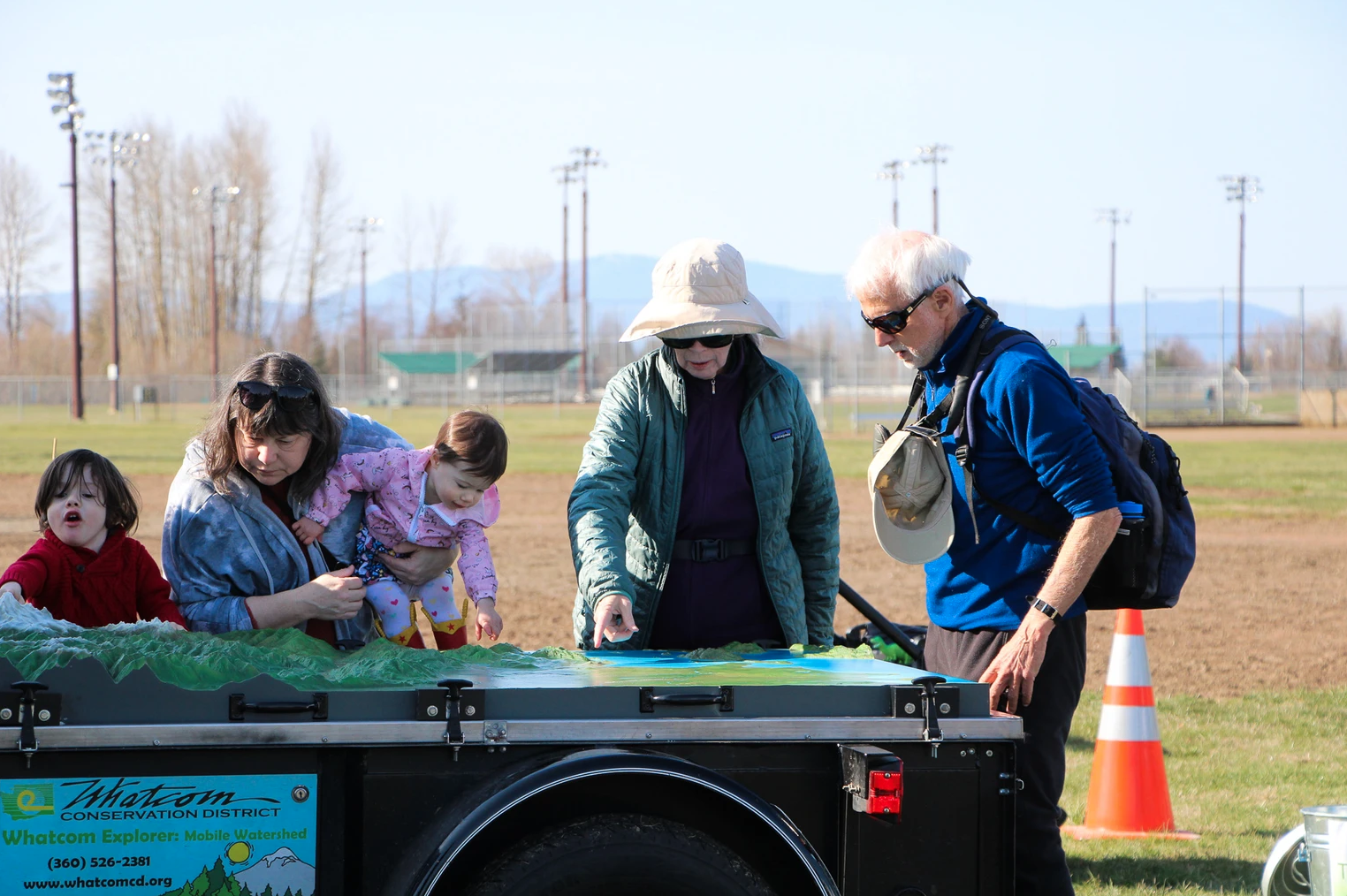 People gathered around an outdoor display table with a map or model, involving adults and children, in a field setting.