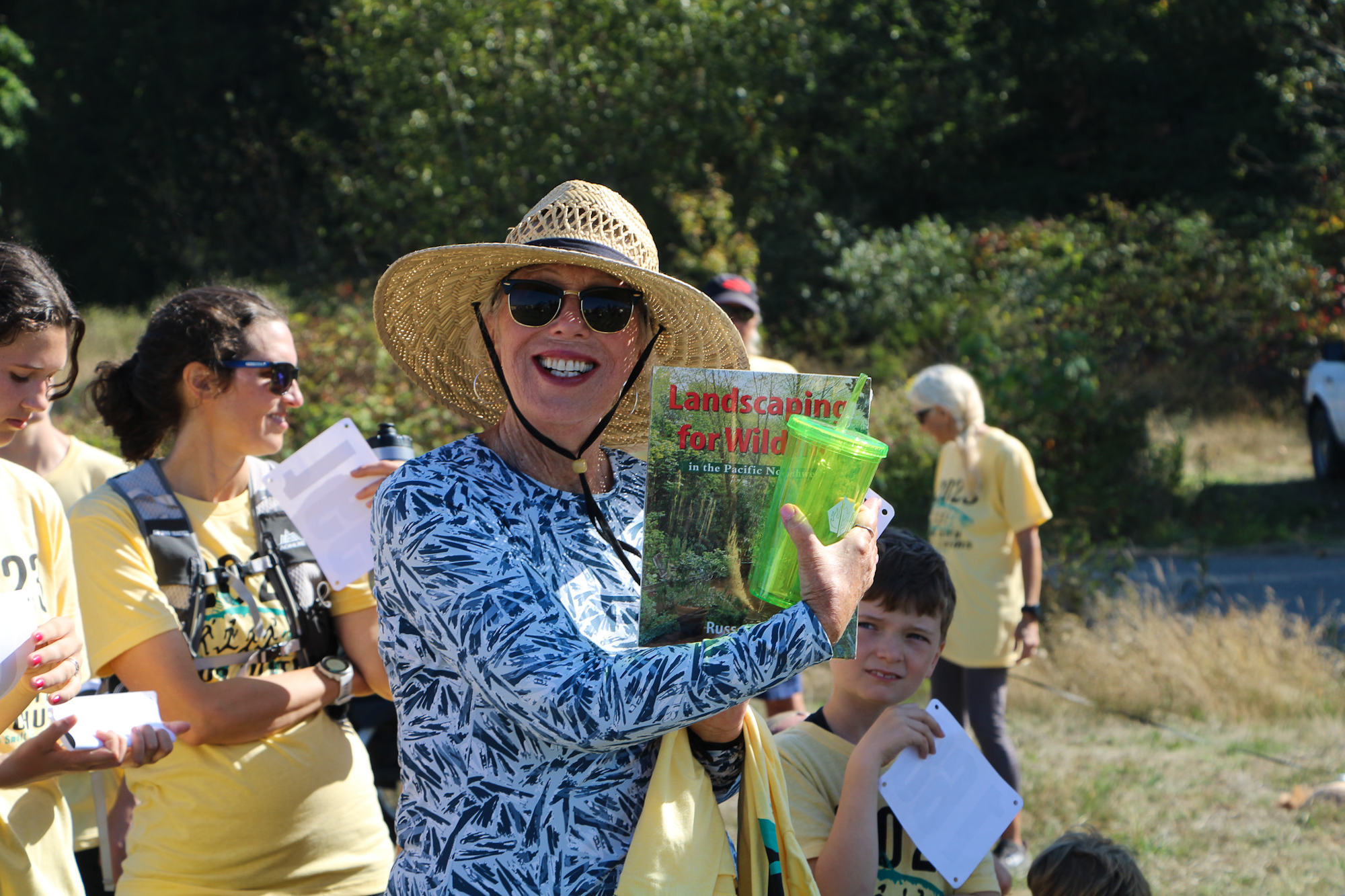 Person in sunglasses and straw hat holding a book titled "Landscaping for Wildlife" and a green cup, surrounded by a group outdoors.