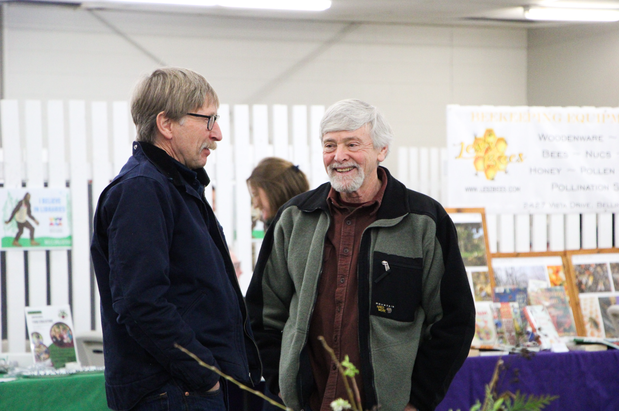 Two men talking and smiling at an indoor event, with informational booths in the background.