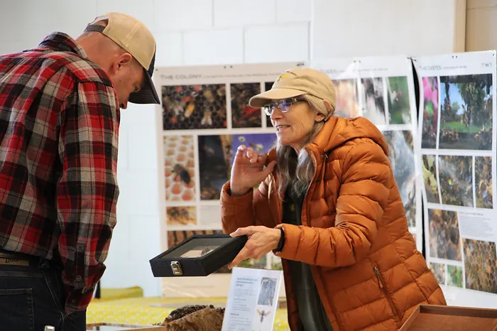 A woman in an orange jacket is enthusiastically showing something in a box to a man in a cap at an exhibit or informational booth.