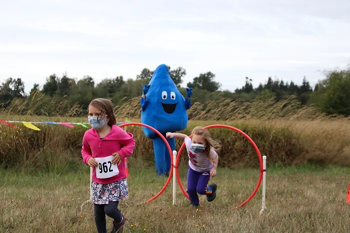 Two kids in a race with a blue mascot in the background, running through hoops in a grassy field.