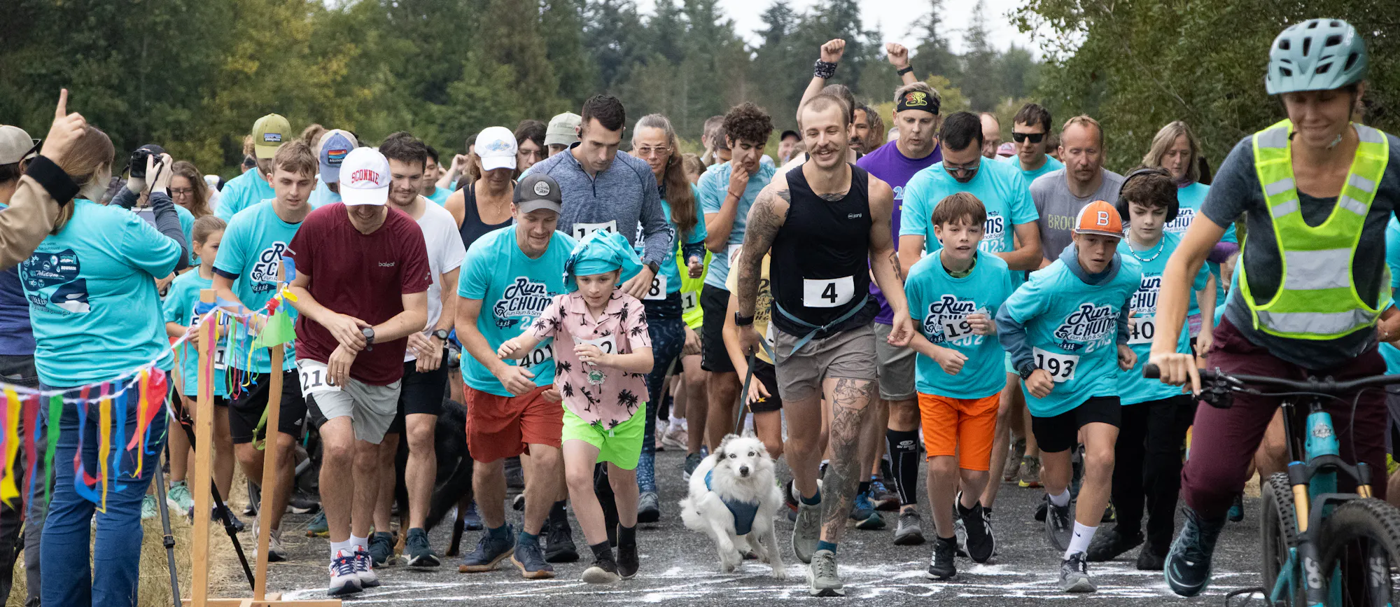 A group of people and a dog are starting a fun run, all wearing blue shirts, amidst a forested backdrop.