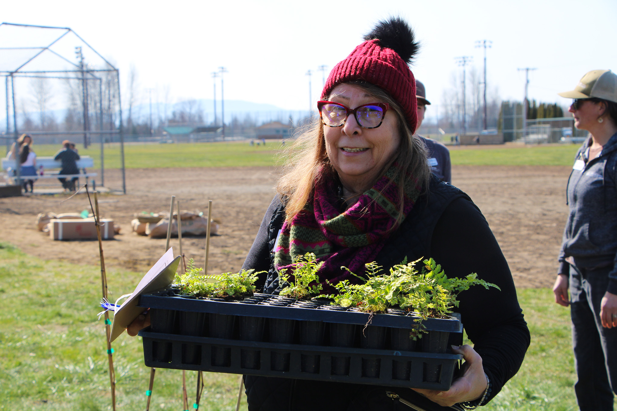 A person wearing a red beanie holds a tray of small plants outdoors, near a field with people in the background.
