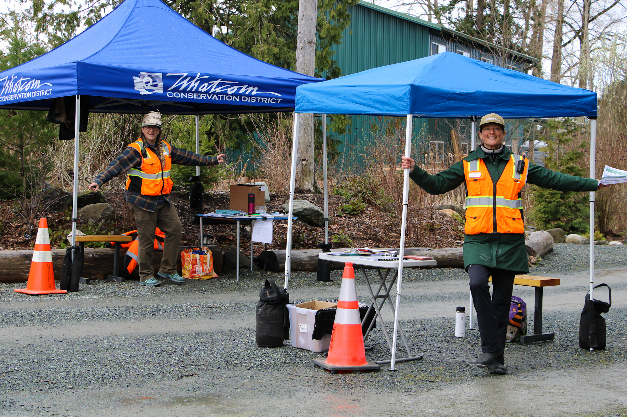 Two people in safety vests under tents at a conservation district event with tables and traffic cones.