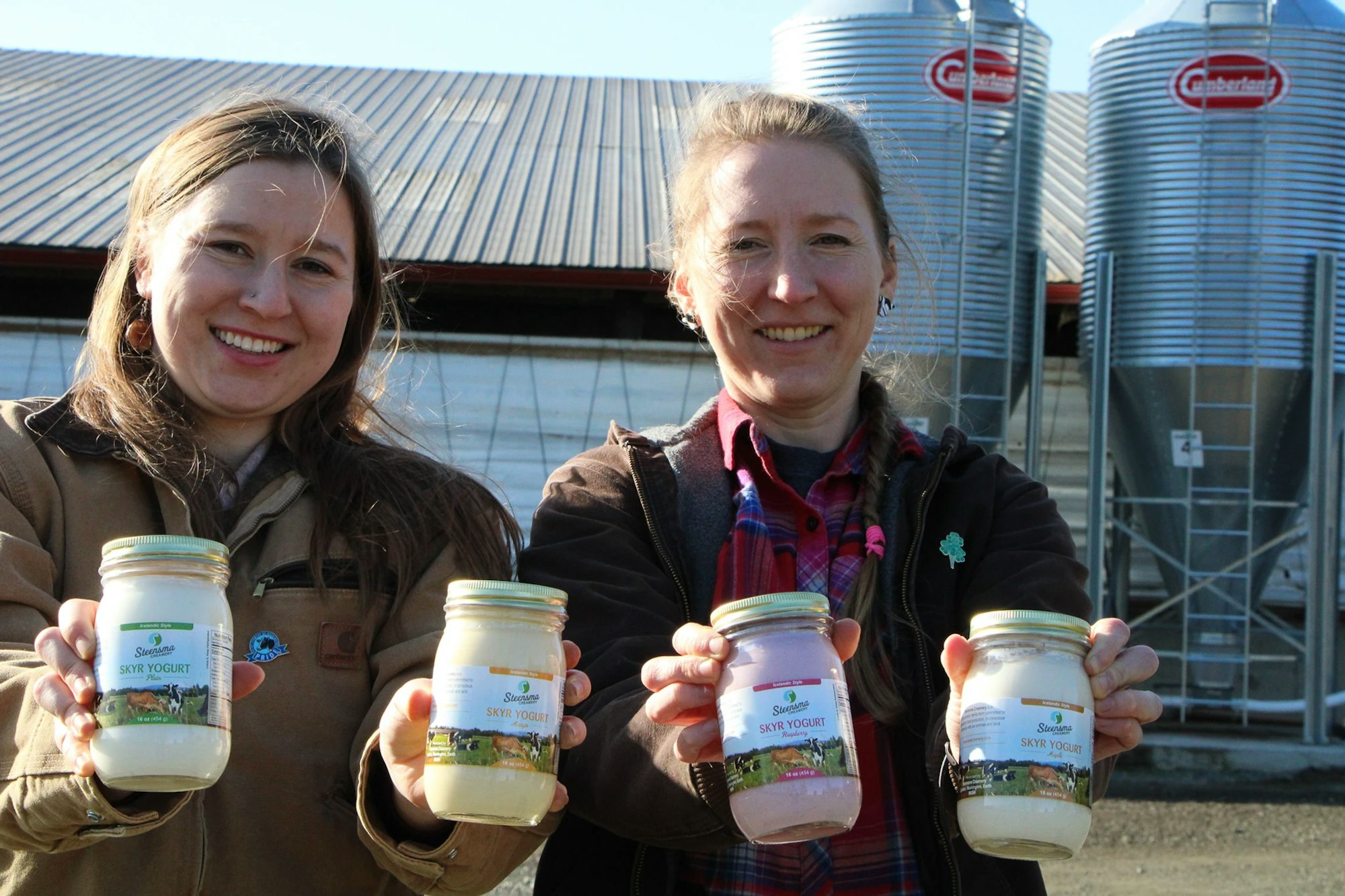 Two people smiling, holding jars of Skyr yogurt in front of a barn and silos.