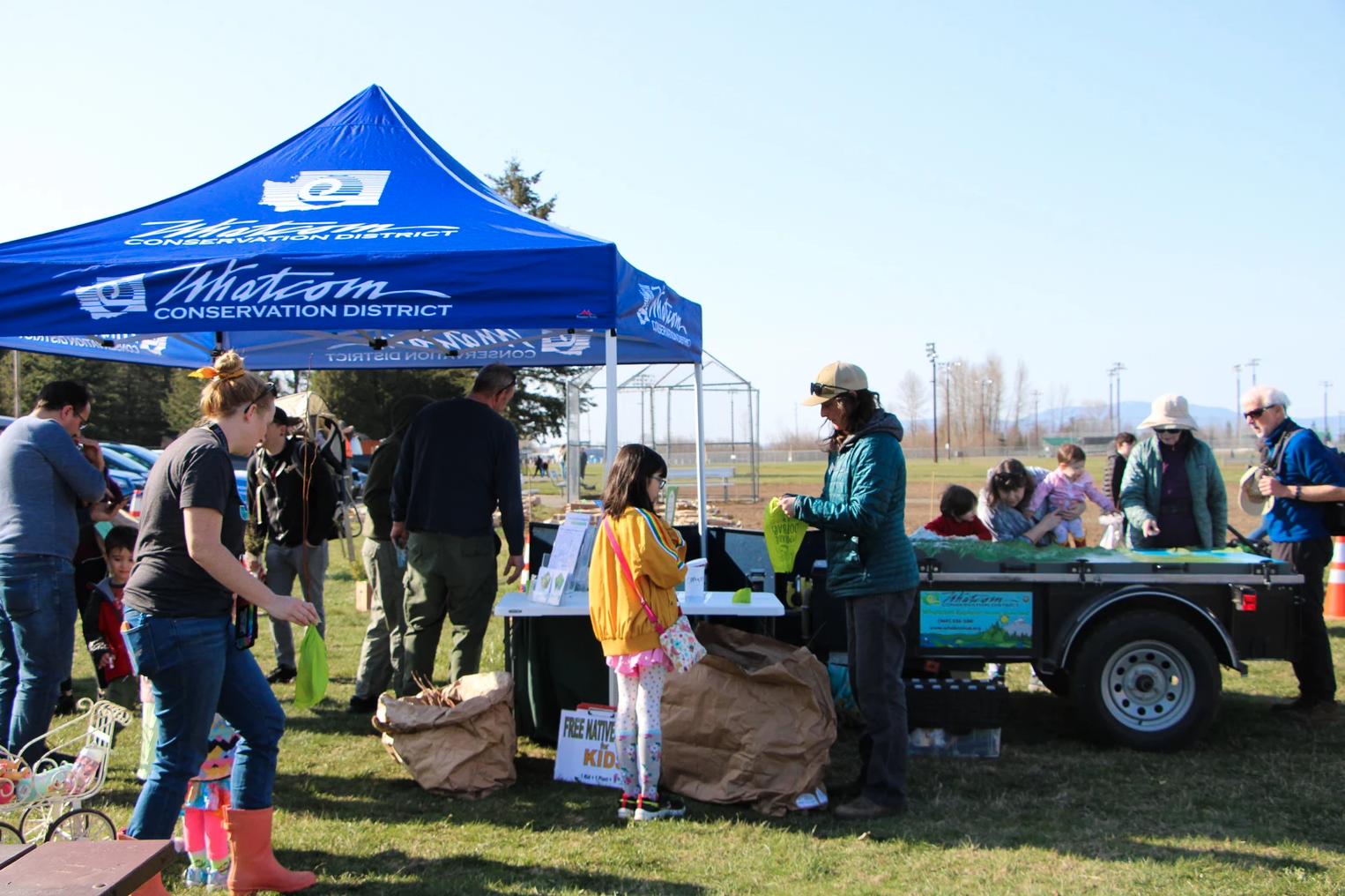 People gathering at a Whatcom Conservation District tent, outdoors, engaging in activities or picking up items.