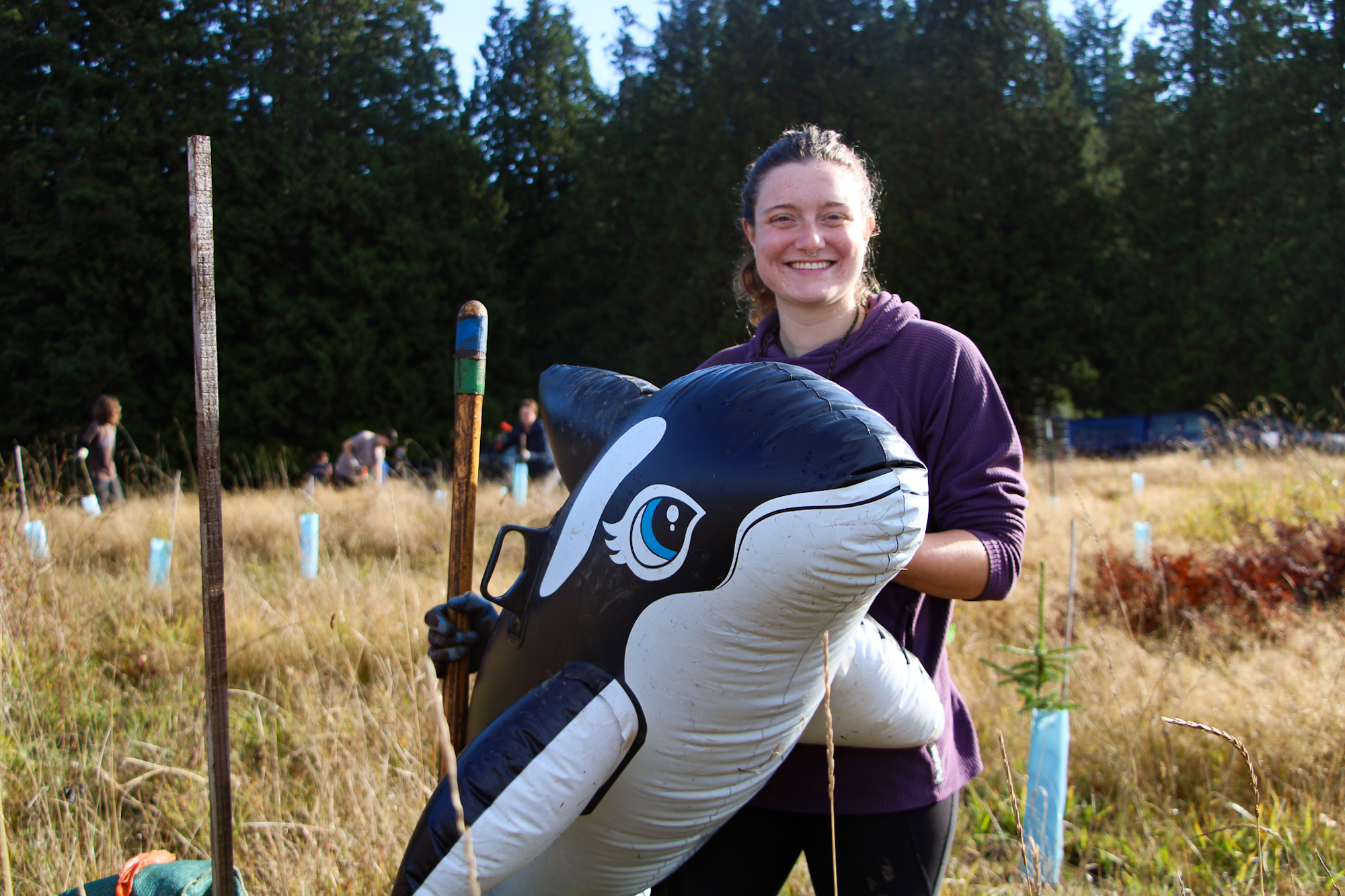 A person in a field holding an inflatable orca, with trees and people working in the background.