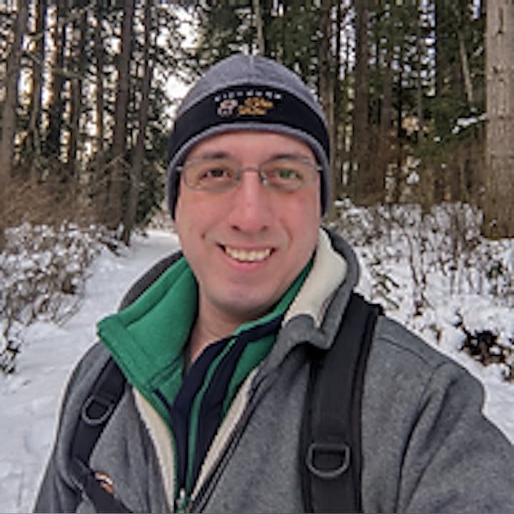 A person wearing winter clothing and a backpack stands on a snowy path in a forest, smiling at the camera.