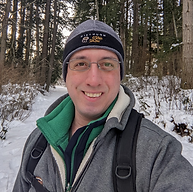 A person wearing winter clothing and a backpack stands on a snowy path in a forest, smiling at the camera.
