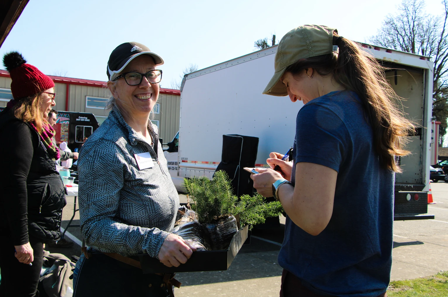 Two people outdoors with plants; one is smiling holding a tray, the other is writing. A trailer and table are in the background.