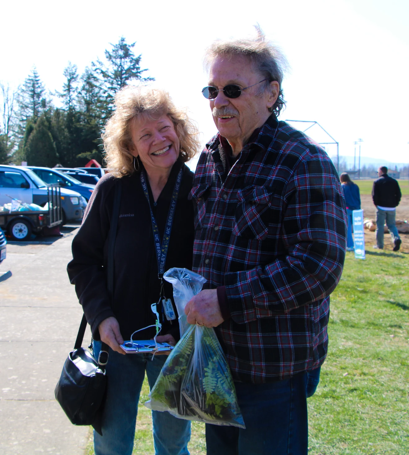 A smiling man and woman are outdoors. He's holding a bag of plants and wearing sunglasses. Cars and people are in the background.