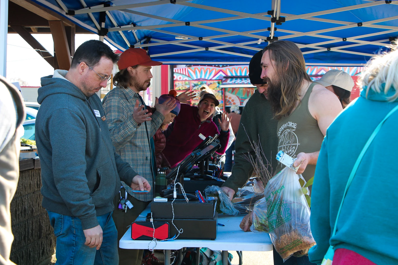 A group of people under a canopy, exchanging goods or plants at an outdoor market or event.