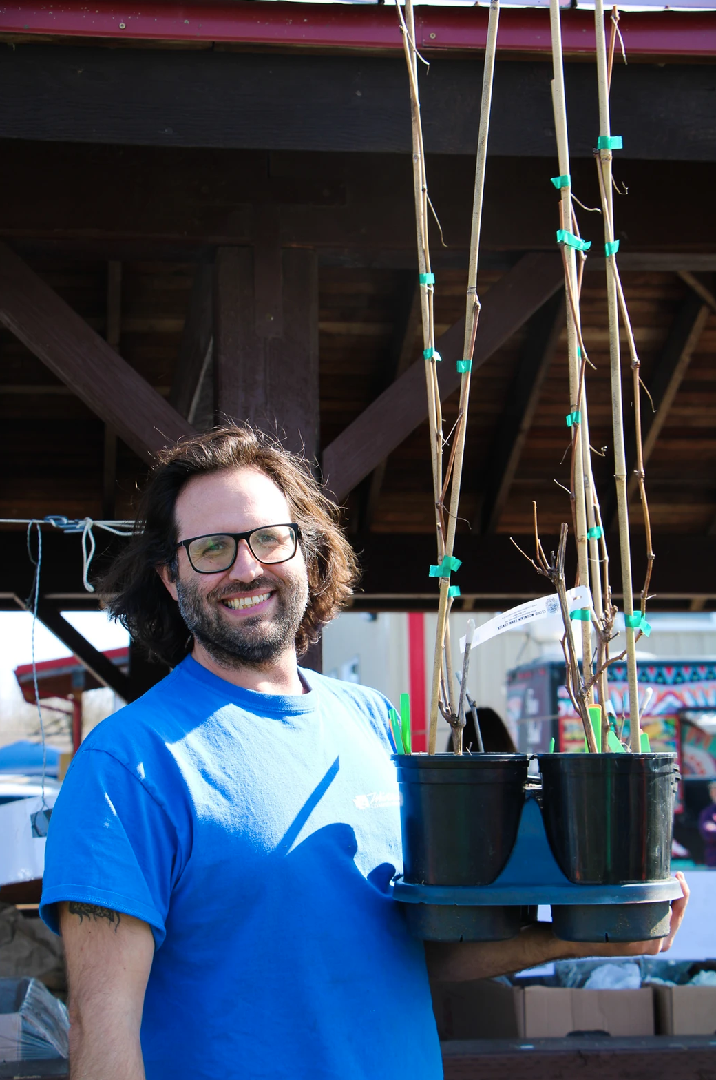 A smiling person holding a tray of potted plants under a wooden structure.