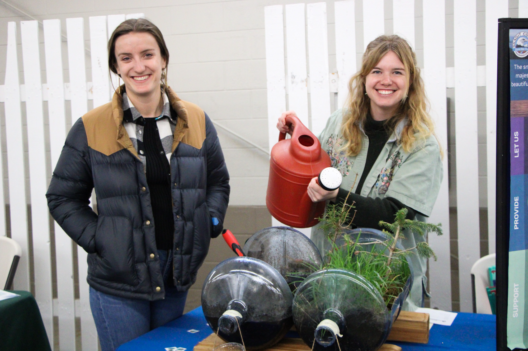 Two people smiling; one holds a red watering can near plants in large bottles on a table.