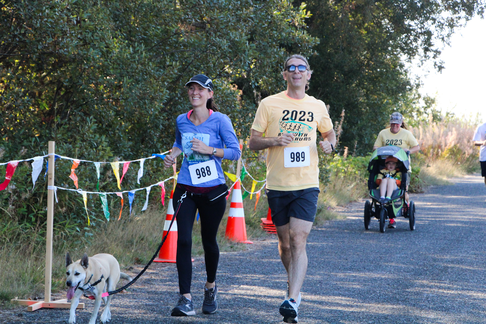 People jogging in a race with dogs and a stroller, wearing numbered bibs, surrounded by trees and colorful flags.