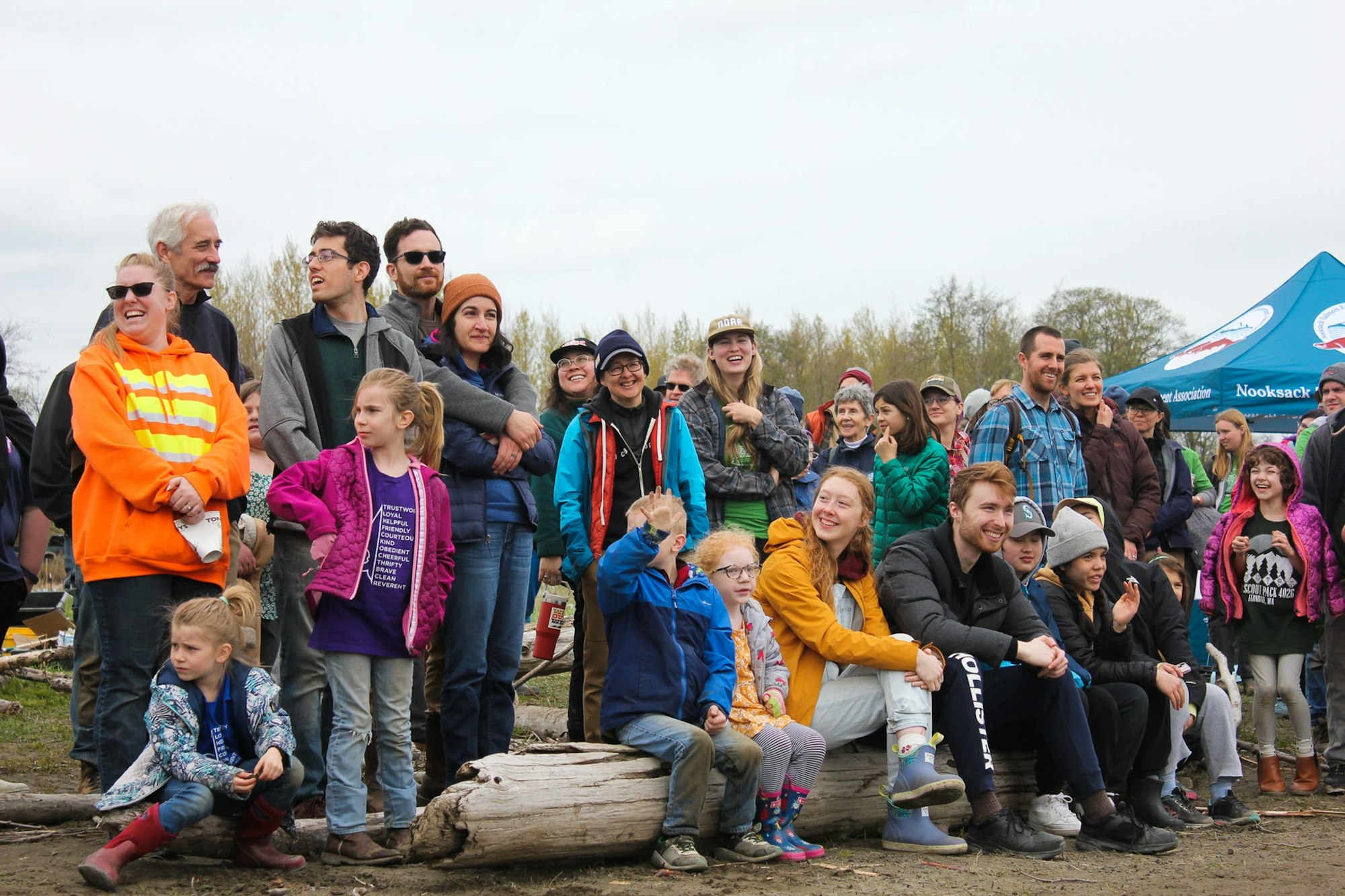 A group of people, including adults and children, gather outdoors on a cloudy day, smiling and looking in one direction.