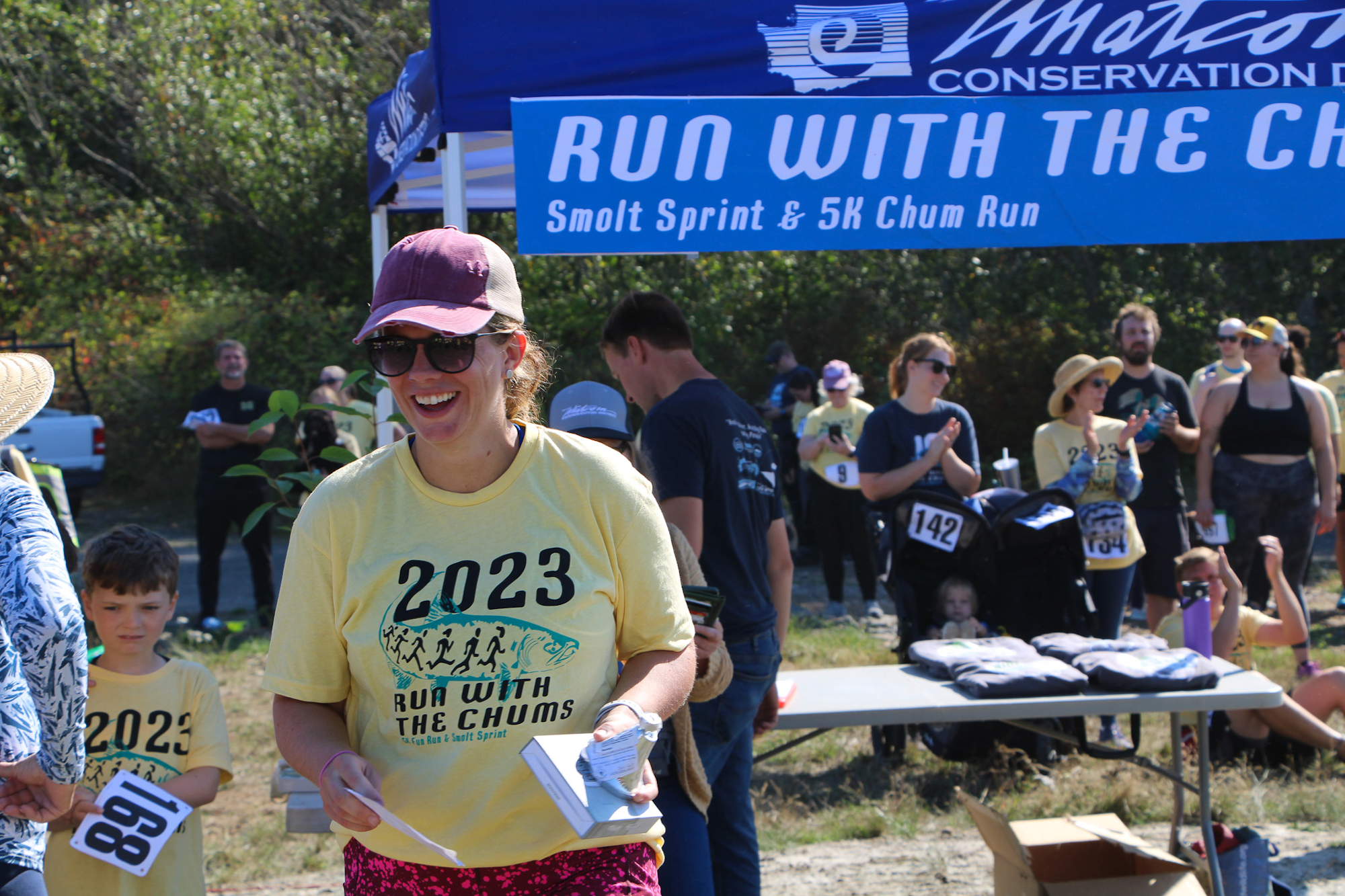 People at an outdoor event with "Run with the Chums" shirts, under a tent. There's a table with items and race bibs visible.