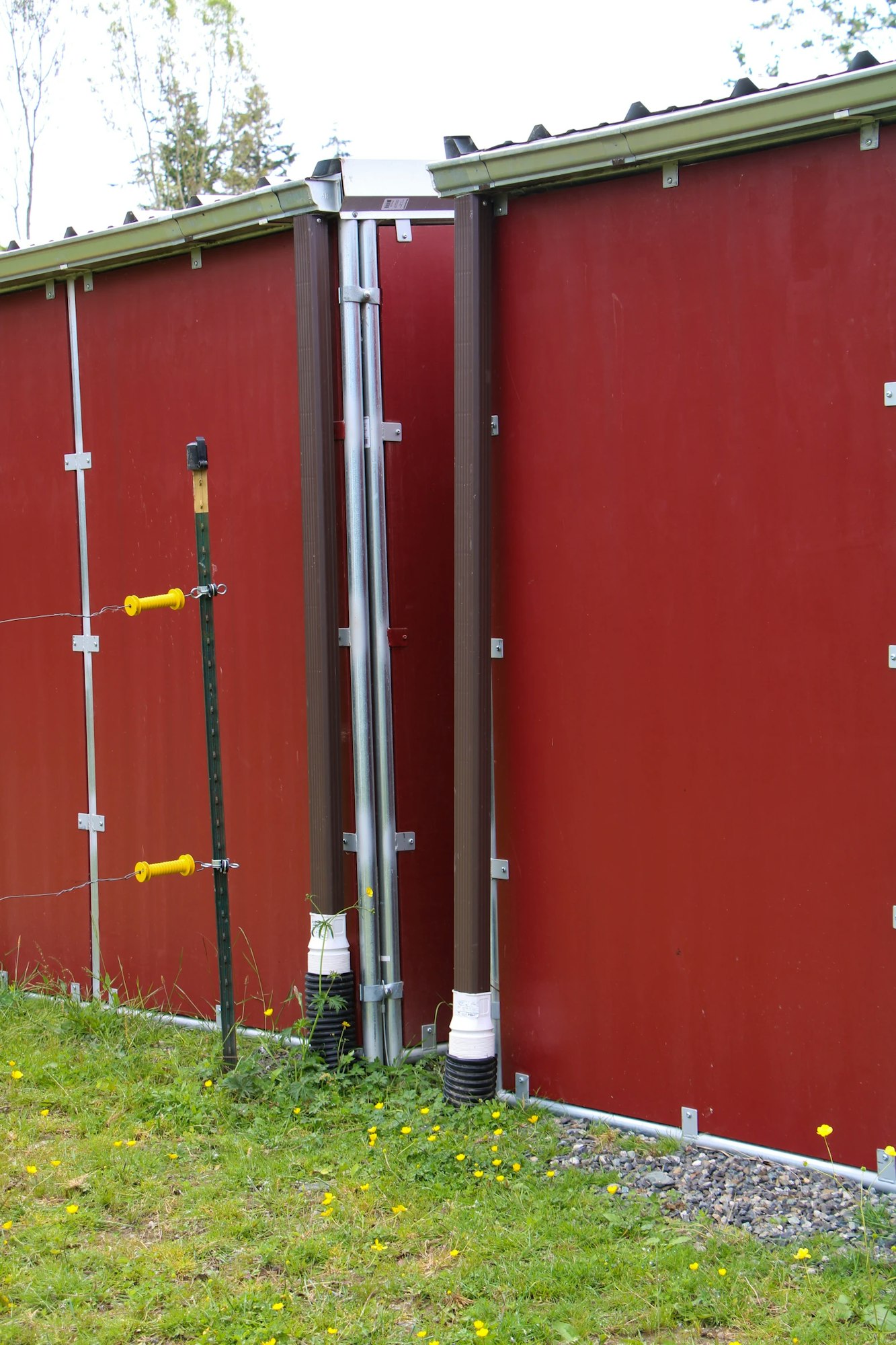 A red wall with metal brackets, pipes, and a door. The ground has grass and yellow flowers.