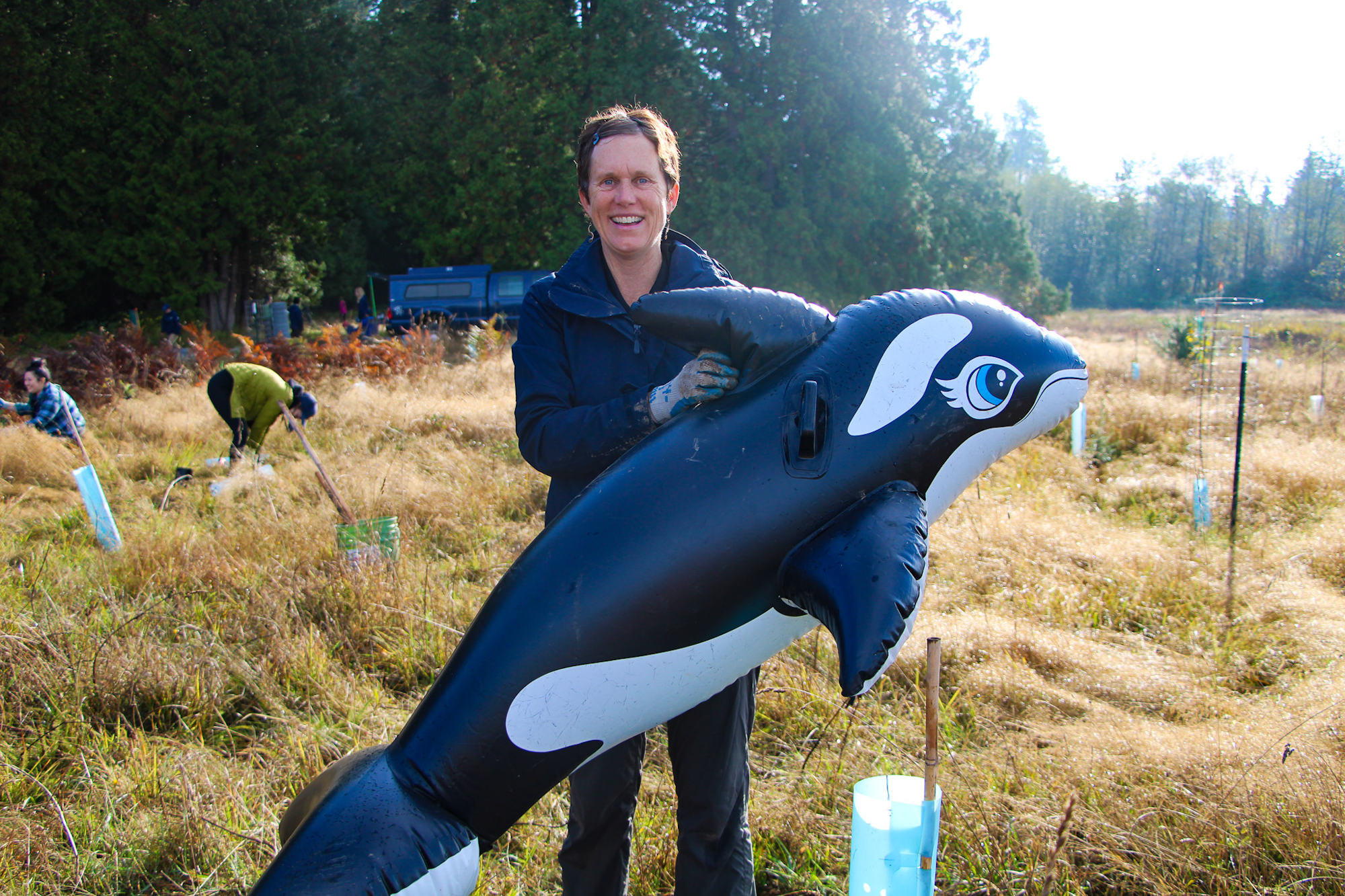 A person in a field holds an inflatable orca, with others working in the background.