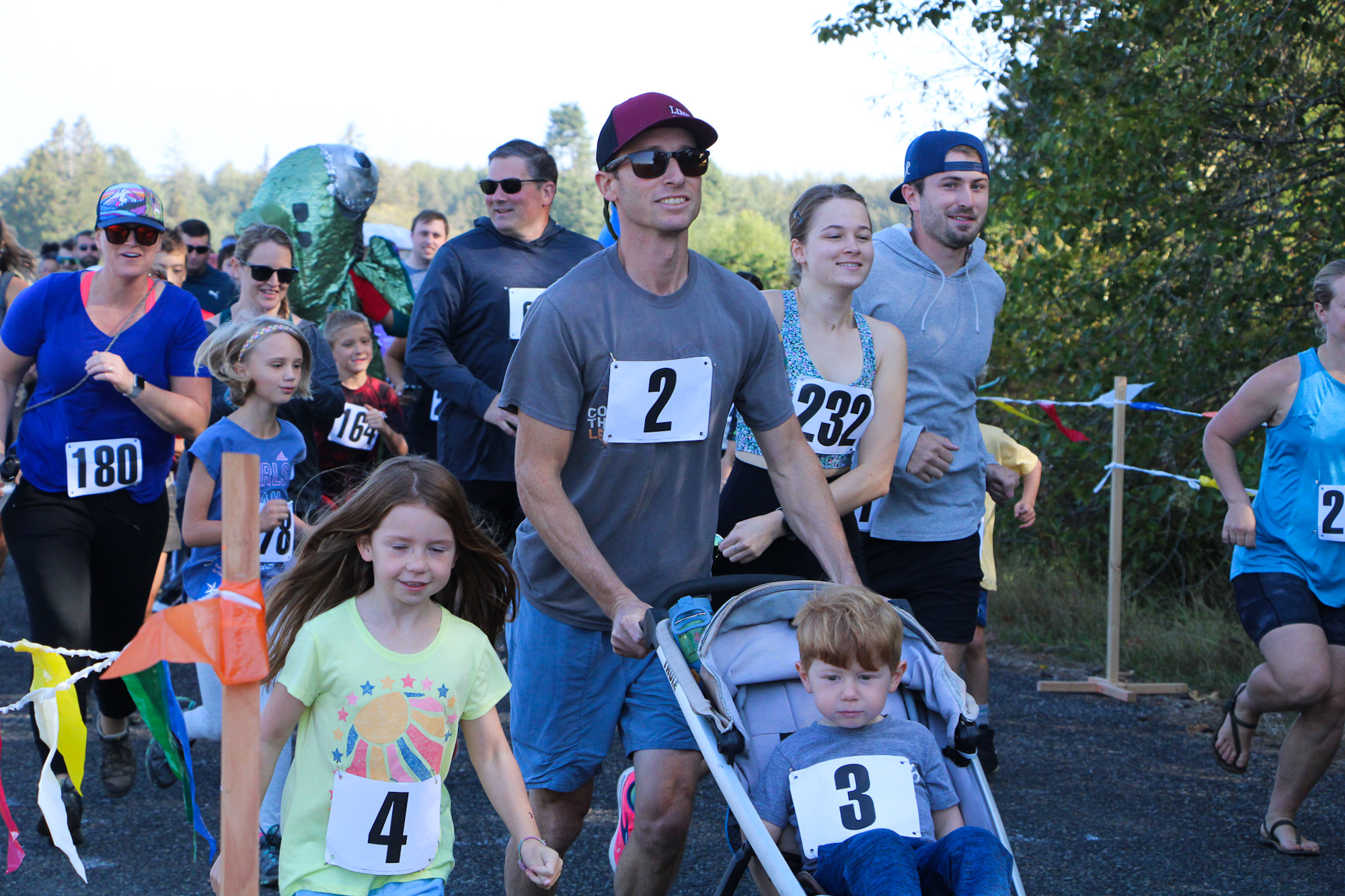 A group of people, including children, running in a race, wearing numbered bibs. One person is pushing a stroller.