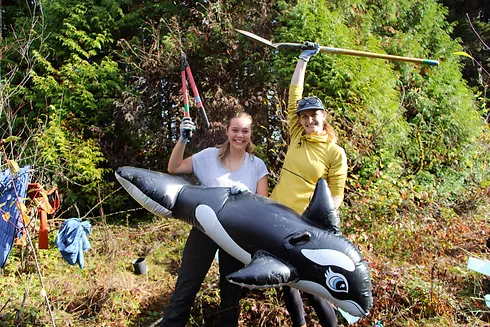 Two people outdoors holding garden tools and an inflatable orca, smiling with greenery in the background.