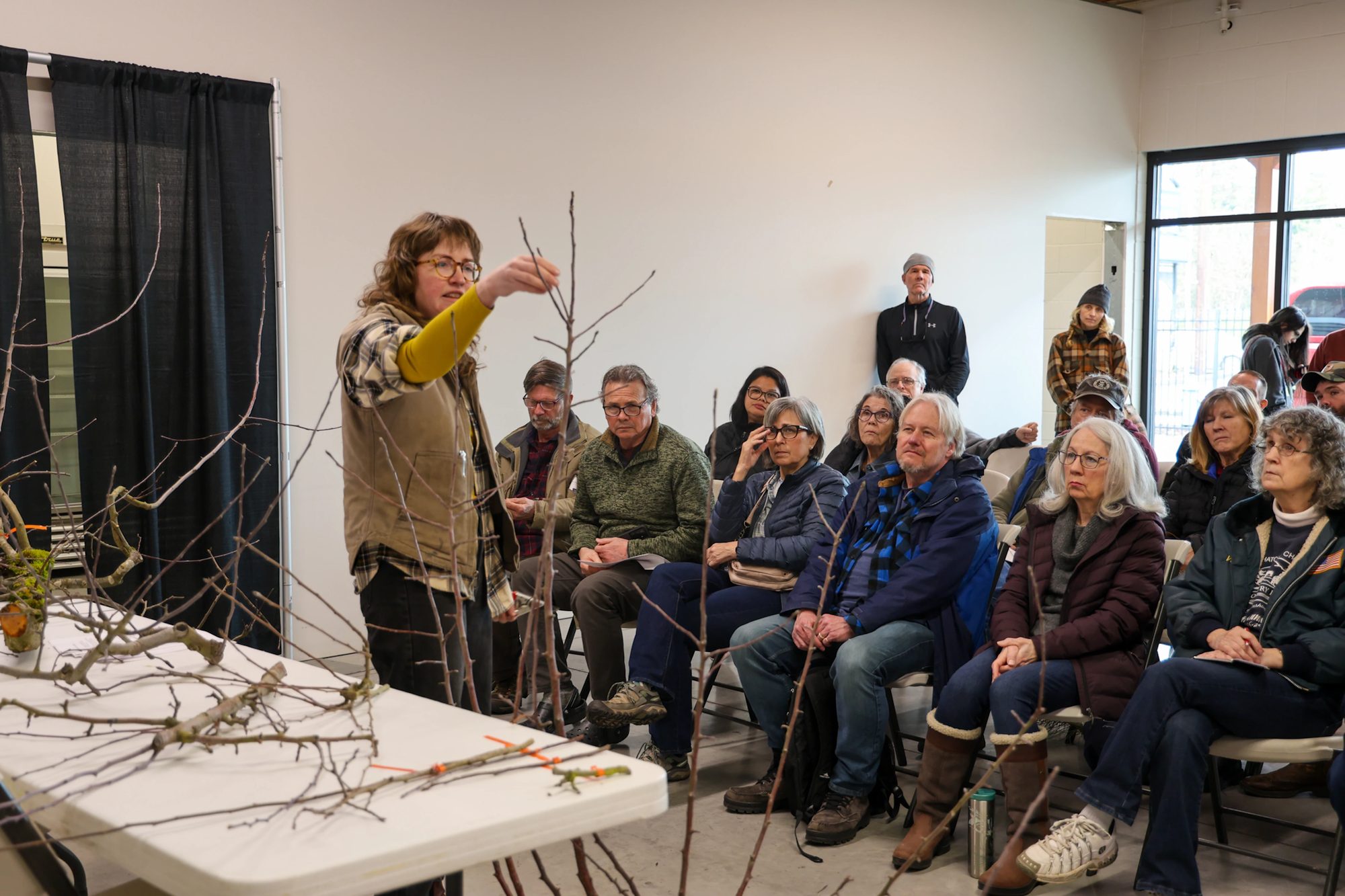 A person teaches a group about tree branches, using samples on a table, in a classroom setting.