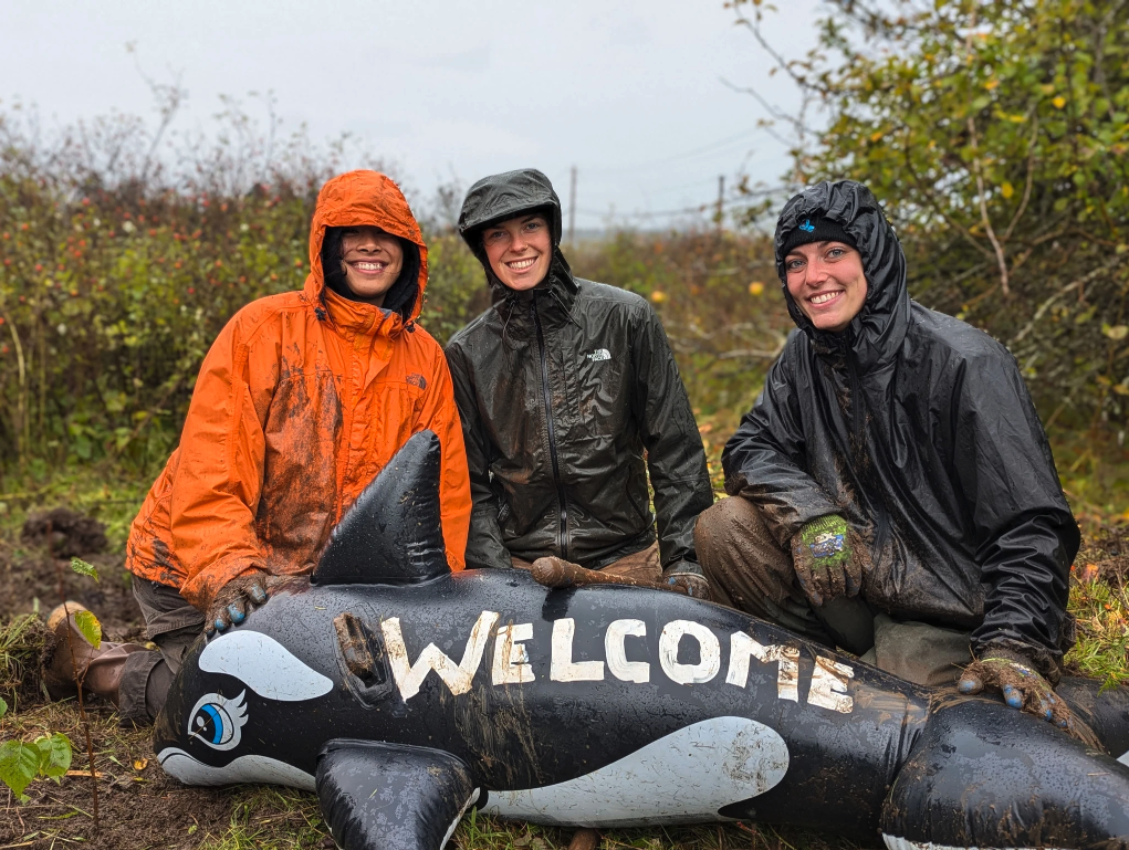Three people in raincoats pose with a "Welcome" inflatable whale, outdoors.