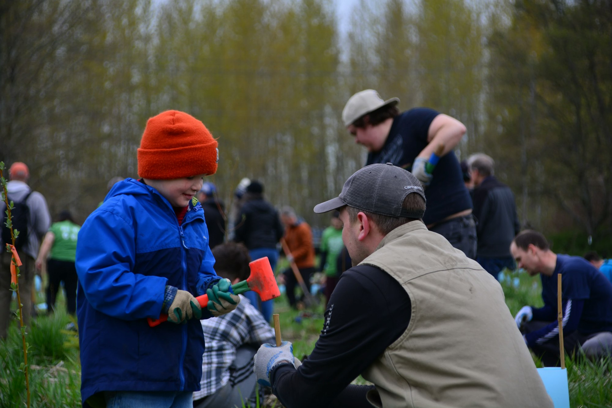 People participating in an outdoor planting activity; a child with a tool and a man kneeling are in the foreground.