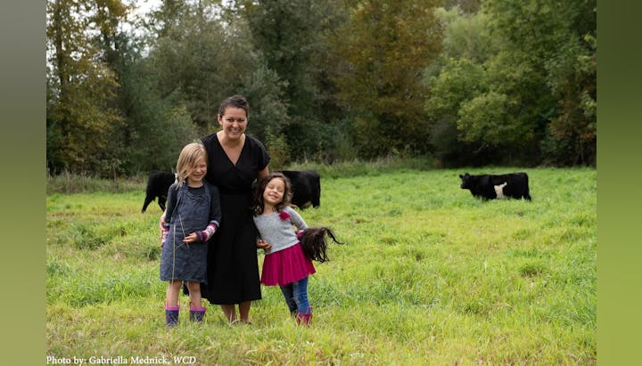 Three people smiling in a grassy field with cows in the background.