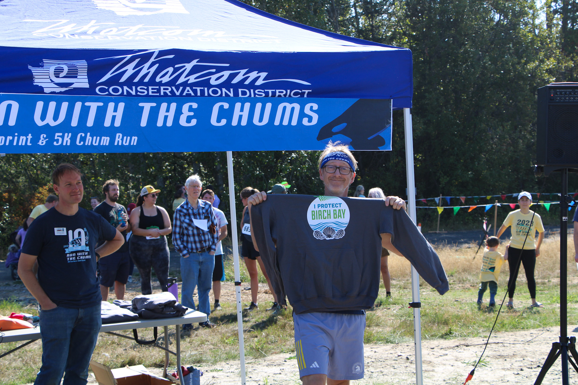 A person holds a "Protect Birch Bay" shirt under a blue tent at an outdoor event with people in the background.